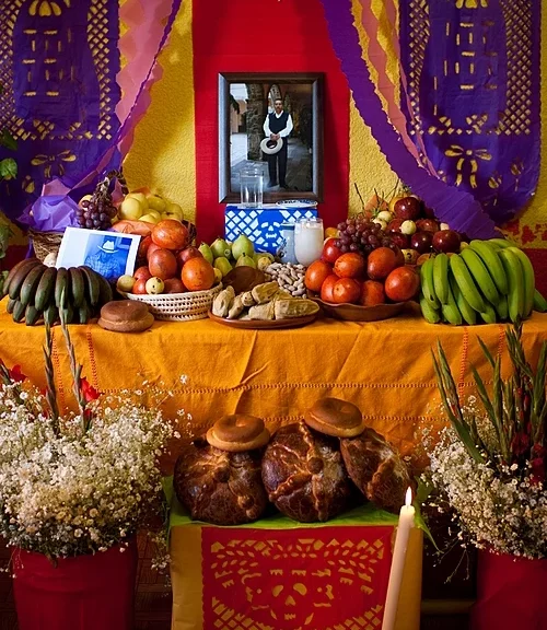 Fotografía de un altar del día de los muertos con cuna mesa con frutas, y comida, y en medio una fotografía del difunto.