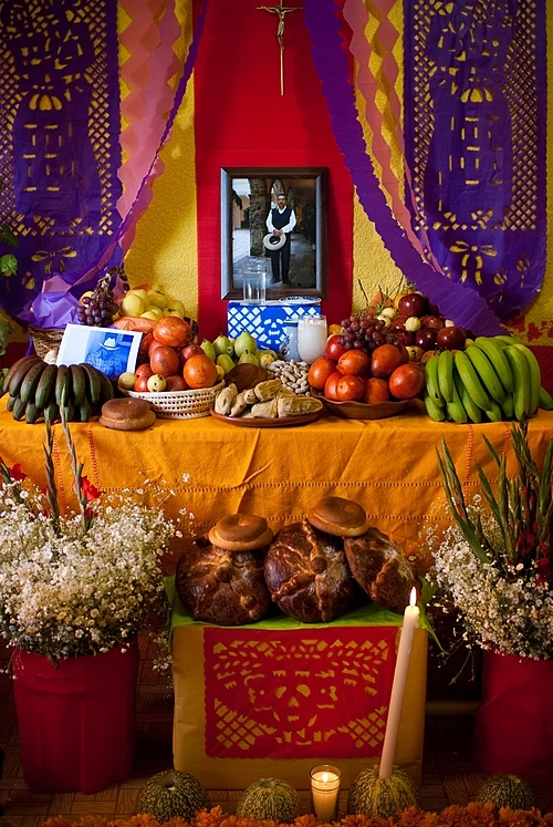 Fotografía de un altar del día de los muertos con cuna mesa con frutas, y comida, y en medio una fotografía del difunto.